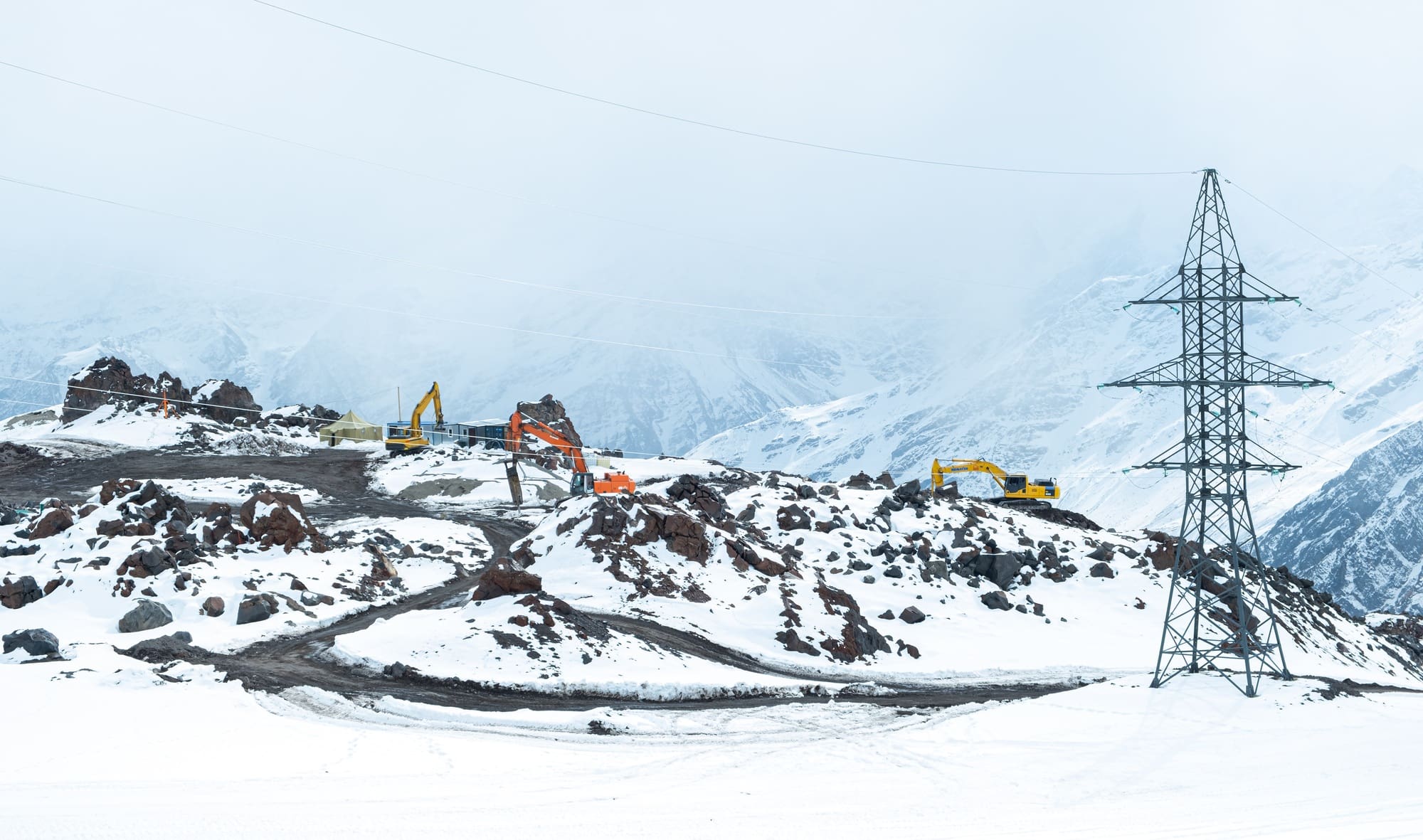 Three excavators are working in the snowy mountains next to the power line.